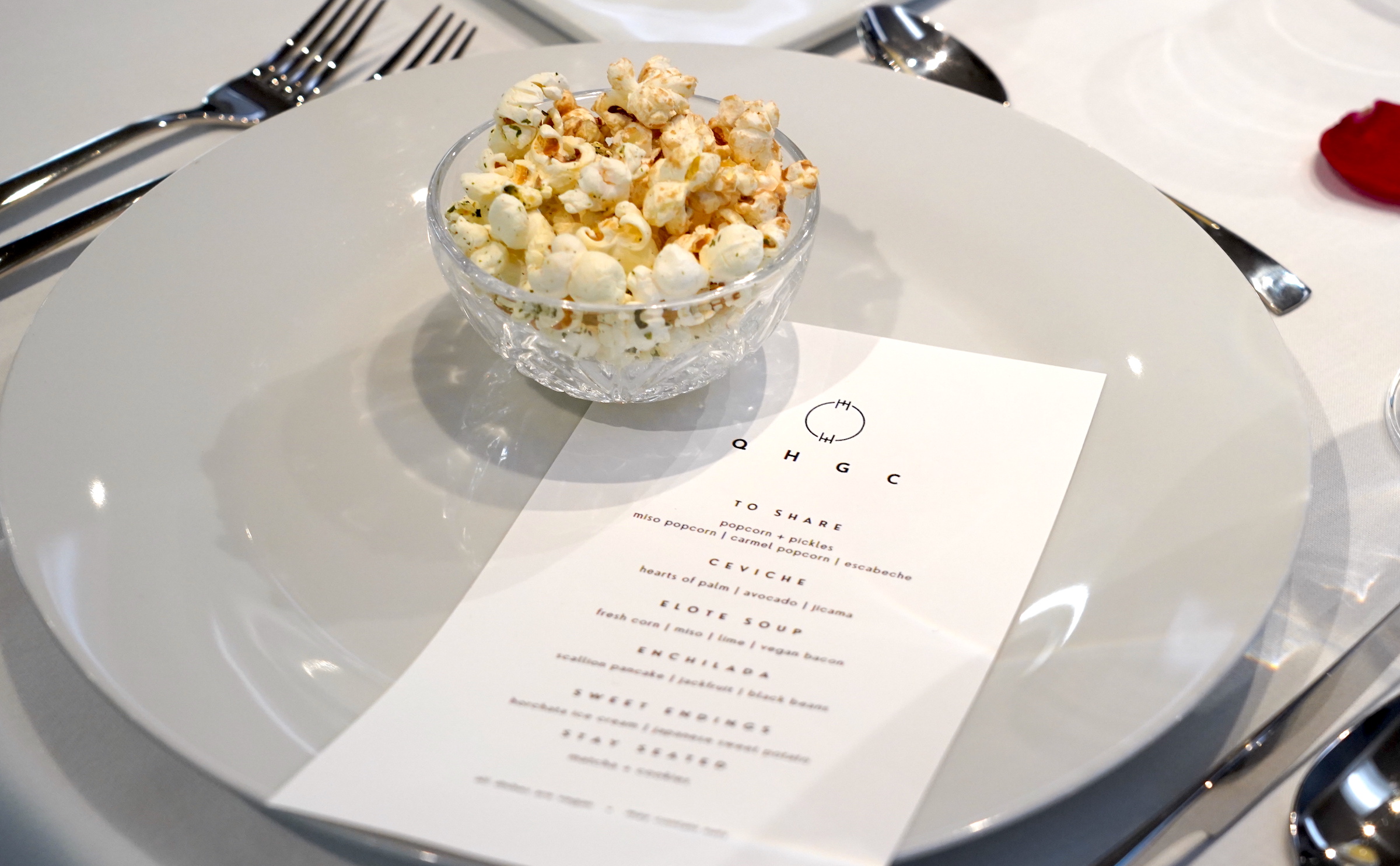 A small glass bowl of popcorn sits on a white plate with a white menu titled "QHGC"