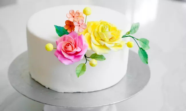A white cake with colorful sugar flower decorations on a silver stand