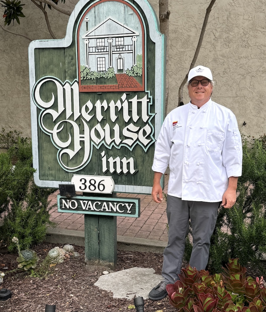Online culinary school graduate Jason Sims smiles in uniform next to the sign for his hotel