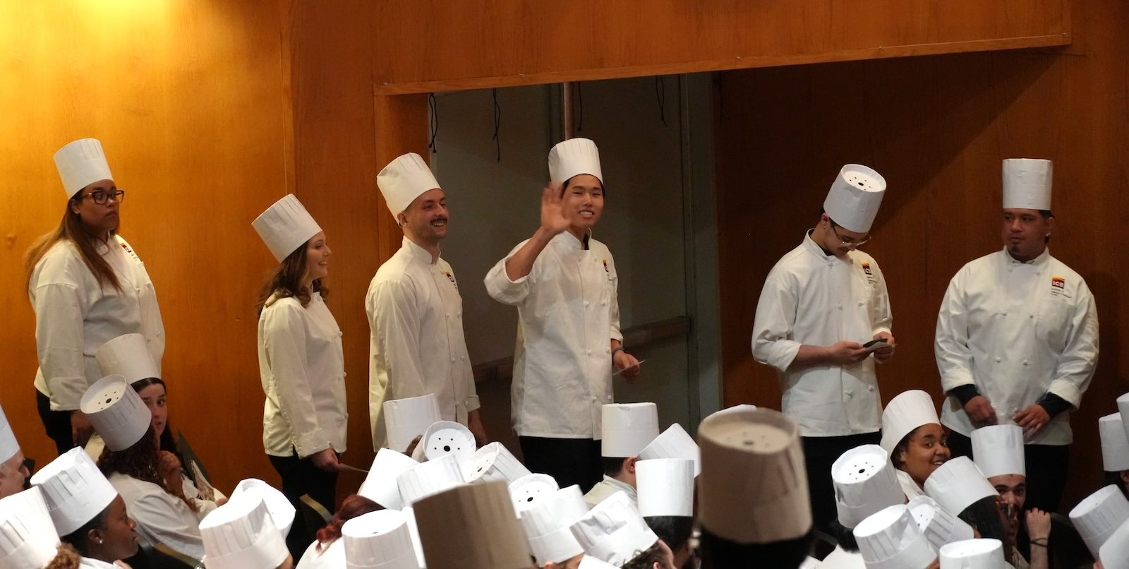 ICE graduate Steven Gao, wearing a white chef's coat and hat, waves while in a line with other graduates at ICE's commencement ceremony