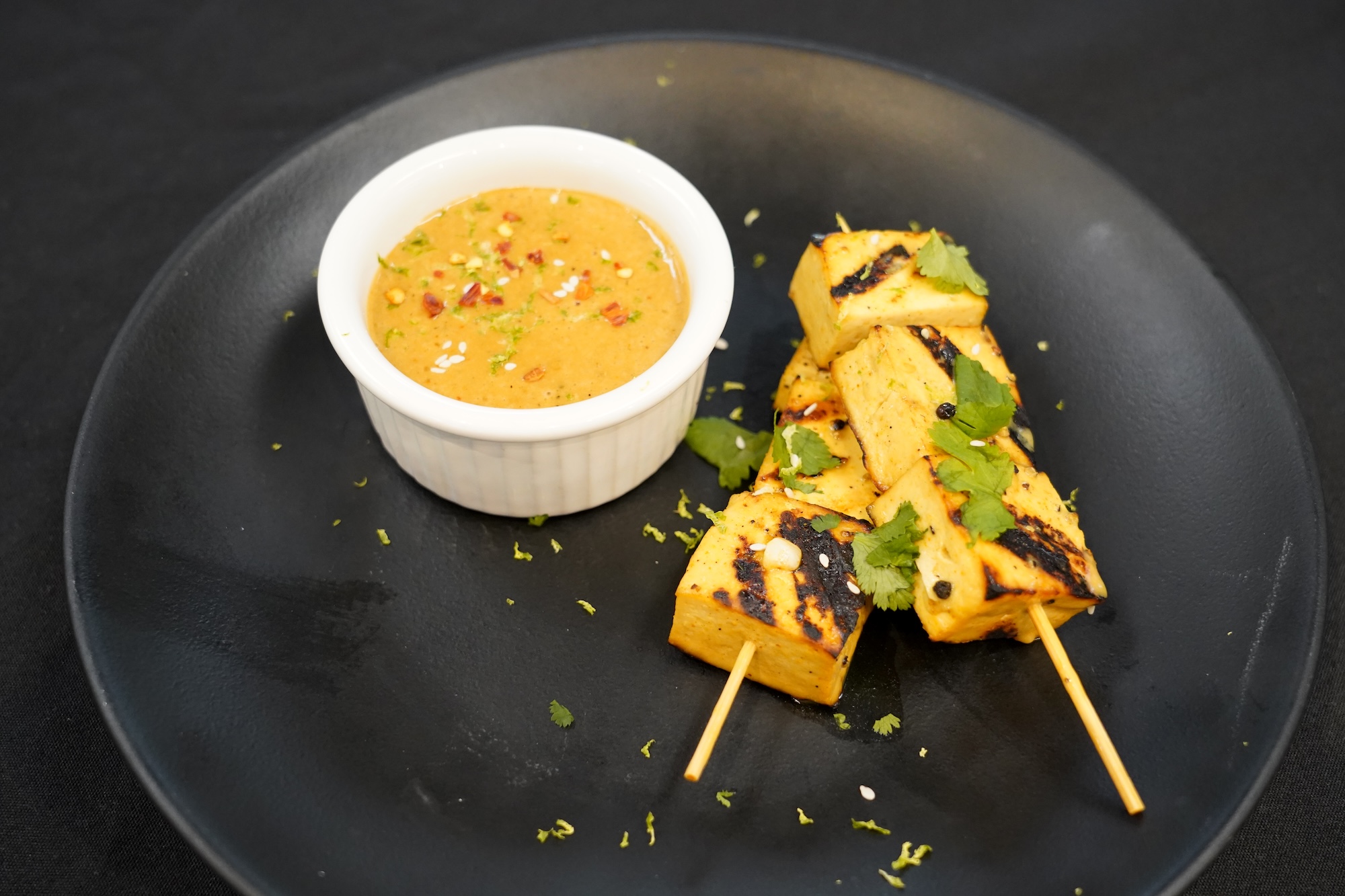 Two tofu skewers with grill marks sit on a black plate next to a white ramekin of tan peanut sauce