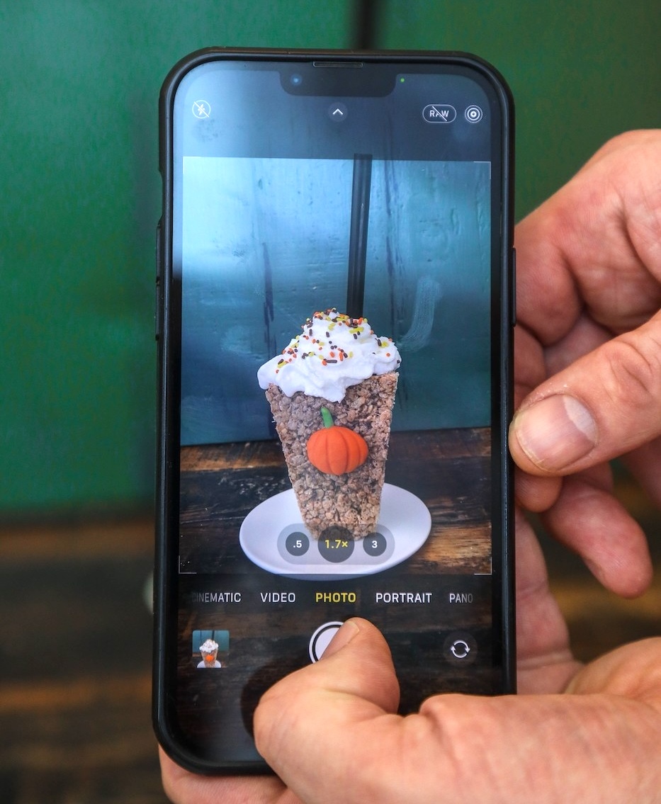 Hands holding an iPhone take a photo of a crispy rice treat shaped to look like a coffee cup with a chocolate pumpkin on it