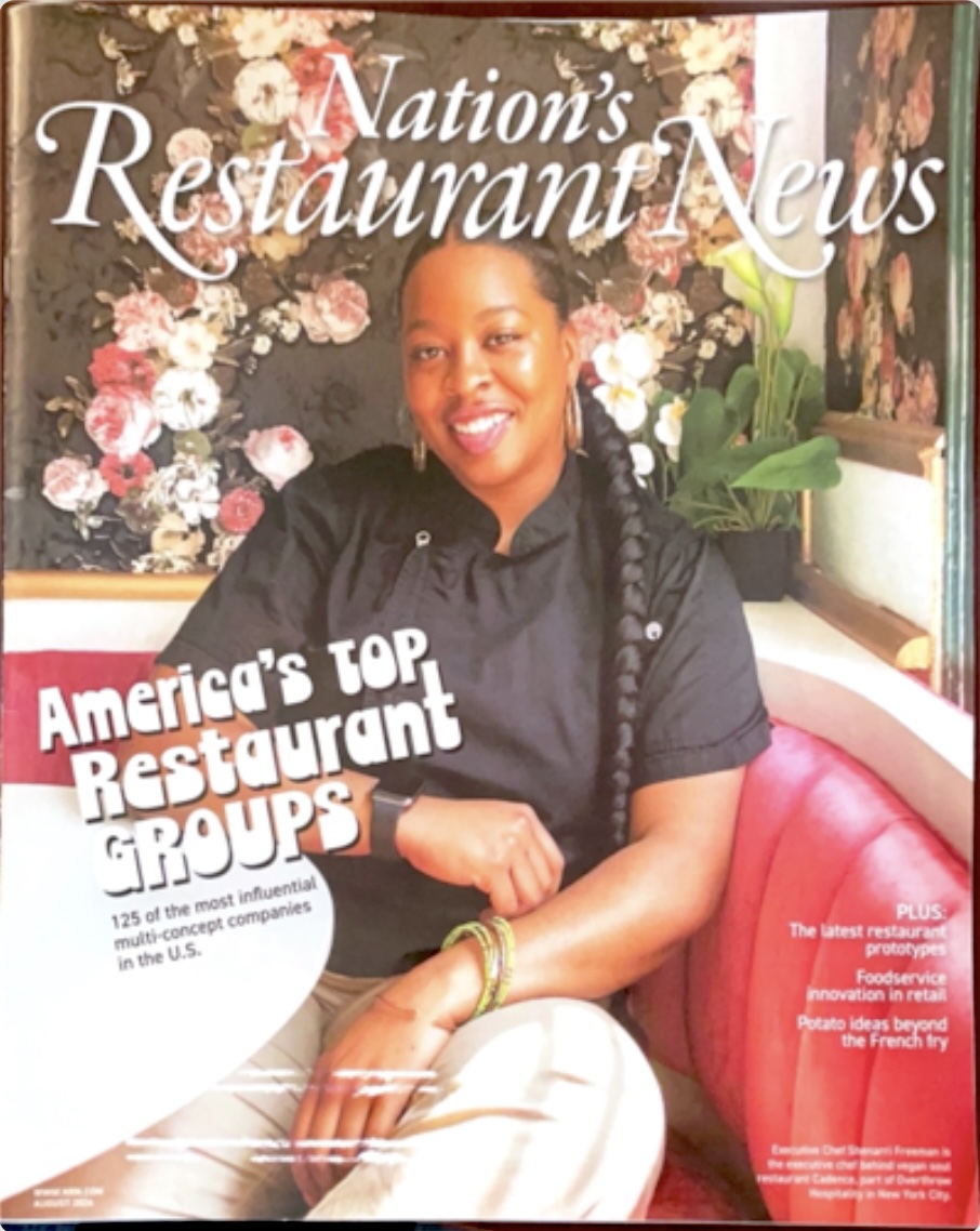 ICE graduate Chef Shenarri Freeman, a woman with black hair in a braid wearing a black chef's coat, smiles in front of a floral wallpaper background