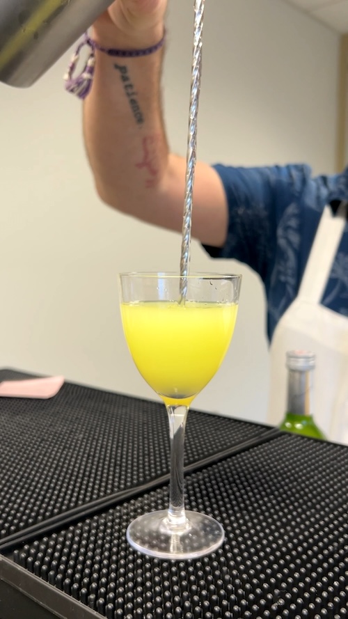 A hand pours water along a metal spoon into a slightly opaque green drink in a Nick and Nora glass