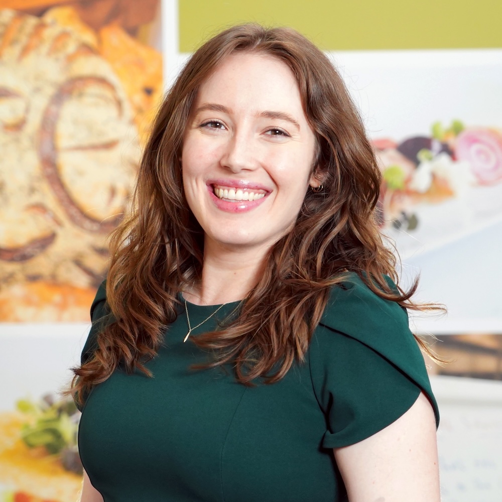 Anna Johnson, a brunette woman in a dark green dress, smiles in front of a wall with pictures of plated food and bread on it