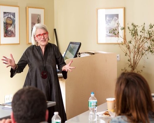 Woman with white hair and bright red glasses wearing all black speaks to Tourism, Travel & Hospitality Management students 