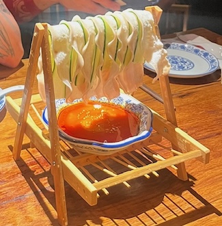 Pork belly and cucumber slices hanging on a wooden bar over sauce