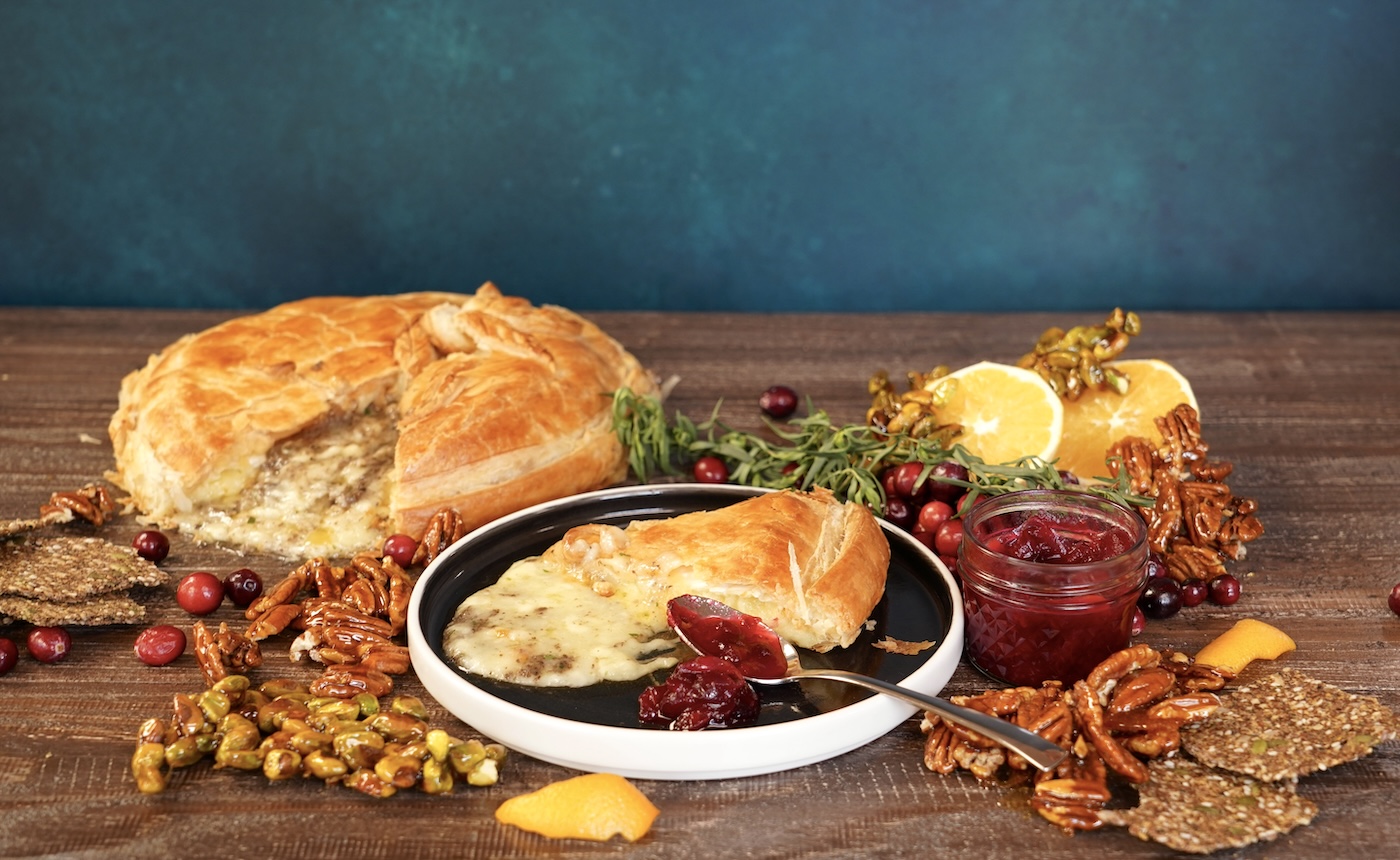 A slice of baked Brie cheese wrapped in baked puff pastry sits on a black plate in front of a wheel of the baked Brie cheese. They are both surrounded by green herbs, orange halves, candied nuts and cranberries