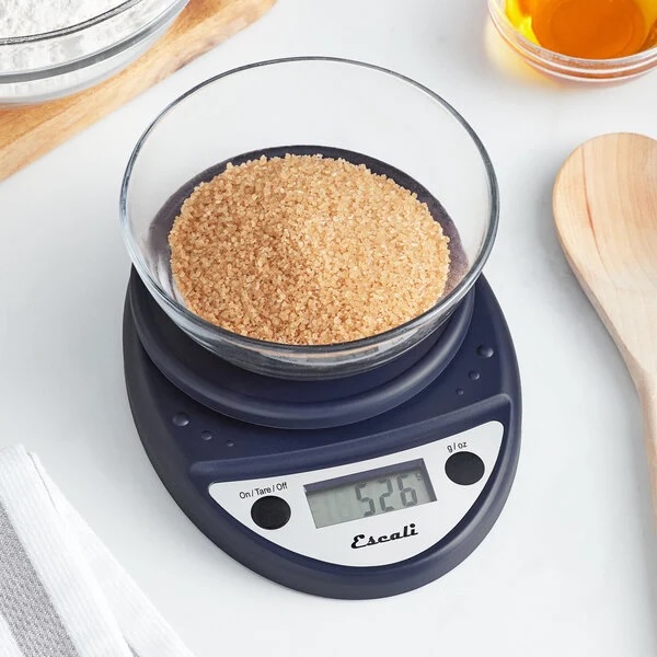 A black kitchen scale sits on a white counter with a glass bowl with brown sugar on top of it