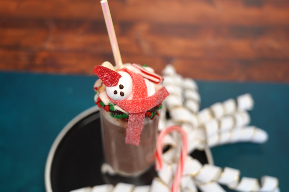 A marshmallow shaped like a snowman with a red gumdrop hat sits on top of a mug of hot chocolate