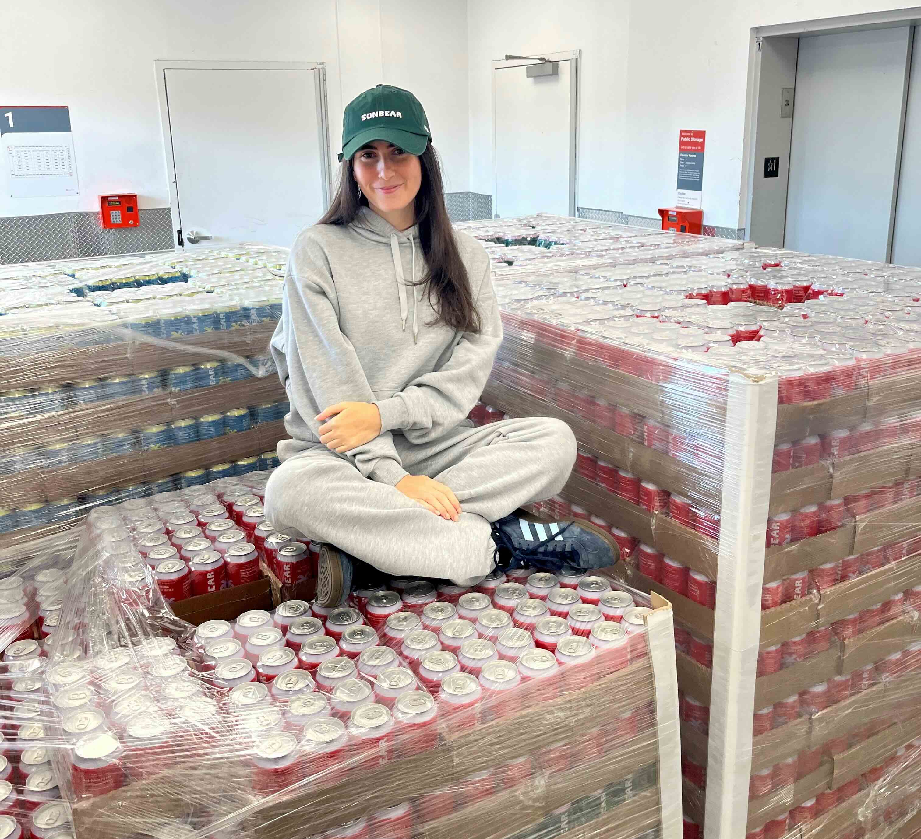 Danielle Krueger, a woman with long brown hair wearing a green hat that reads "Sunbear" and wearing a light grey sweat suit, sits on pallets of cans