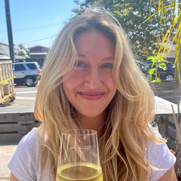 Olivia Jebrine, ICE Culinary Arts Student and contributing food writer to the ICE blog, sits at picnic table toasting with glass of white wine in casual white t-shirt with loosely curled blond hair and smile.