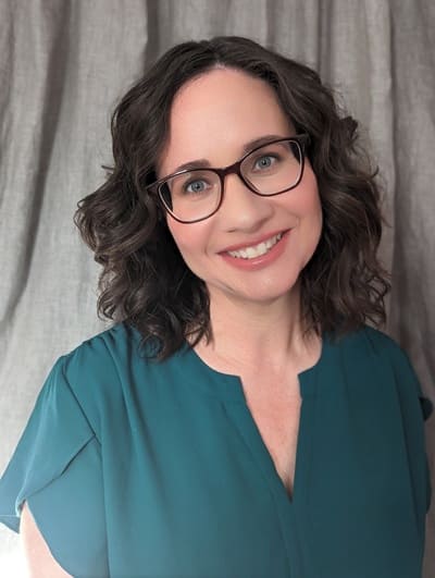 Food writer and cheese expert Pamela Vachon wearing green shirt, wavy brown hair and glasses standing against a curtained backdrop and smiling.