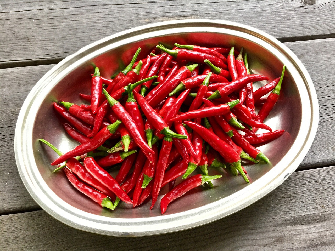 Numerous bright red chili peppers with green stems in a white oval bowl on a wood tabletop.