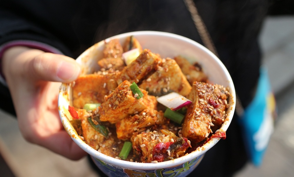 A person's hand holds a bowl filled with sauced pieces of tofu and vegetables