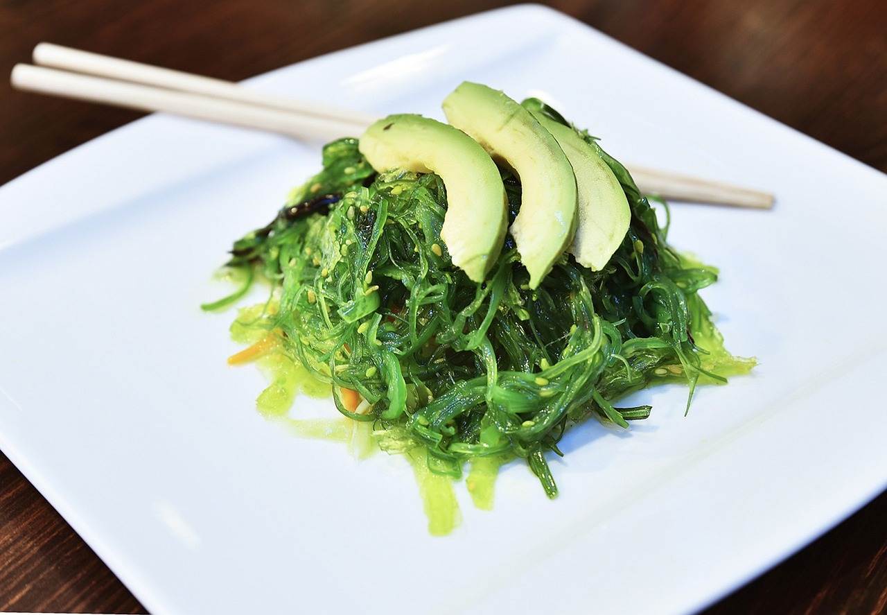 Seaweed salad topped with three slices of avocado sits on white plate with chopsticks beside. 