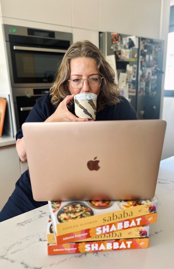 Adeena Sussman sips cofee while working on apple laptop that sits on marble countertop and a stack of her cookbooks titled Shabbat.