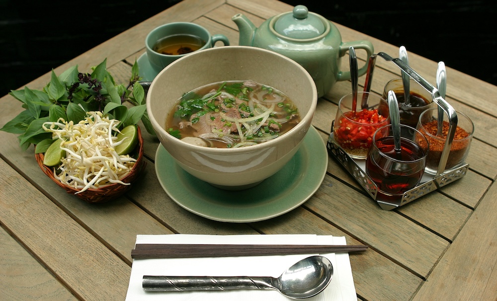 A bowl of pho soup surrounded by sauces, bean sprouts and herbs and a tea kettle in a white bowl on a wooden table