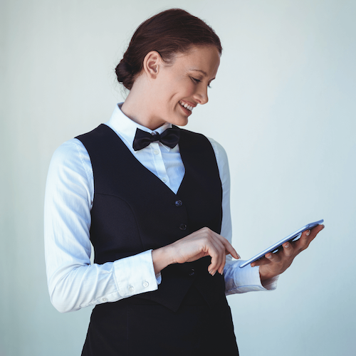 Profile view of a smiling server in a vest and bow tie using a mobile tablet against a plain background.