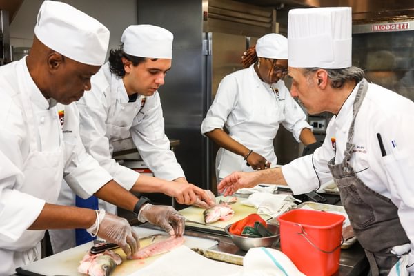 A chef instructor wearing a tall chef's hat guides three culinary students as they practice fish butchery in a commercial training kitchen. The instructor points toward a raw fish on a cutting board while a student examines it closely. In the foreground, another student uses a knife to filet a fish, while a third student works at a prep station in the background.