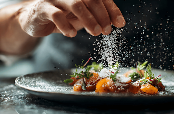 A dramatic, close-up shot of a chef's hand sprinkling fine white powder—such as salt, sugar, or spice—over a gourmet dish. The powder is frozen mid-air, creating a glittering stream under sharp lighting. The dish features small, bright orange pieces of food garnished with fresh green microgreens, all served on a dark, textured plate.