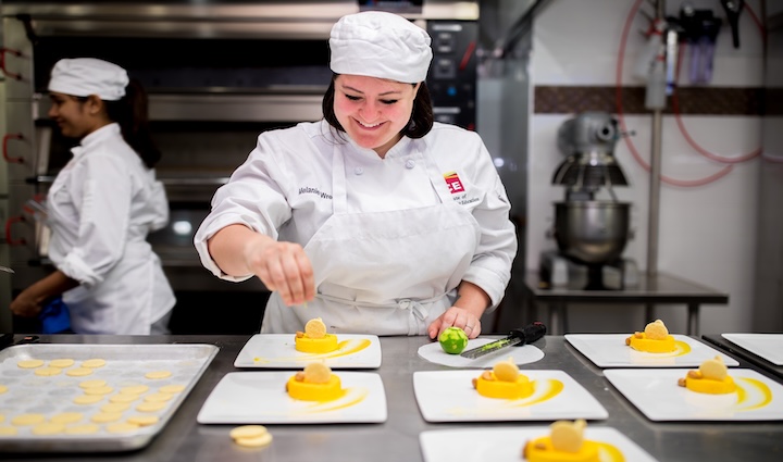 A student in chef's whites smiles while plating vibrant pastry dishes in a kitchen classroom