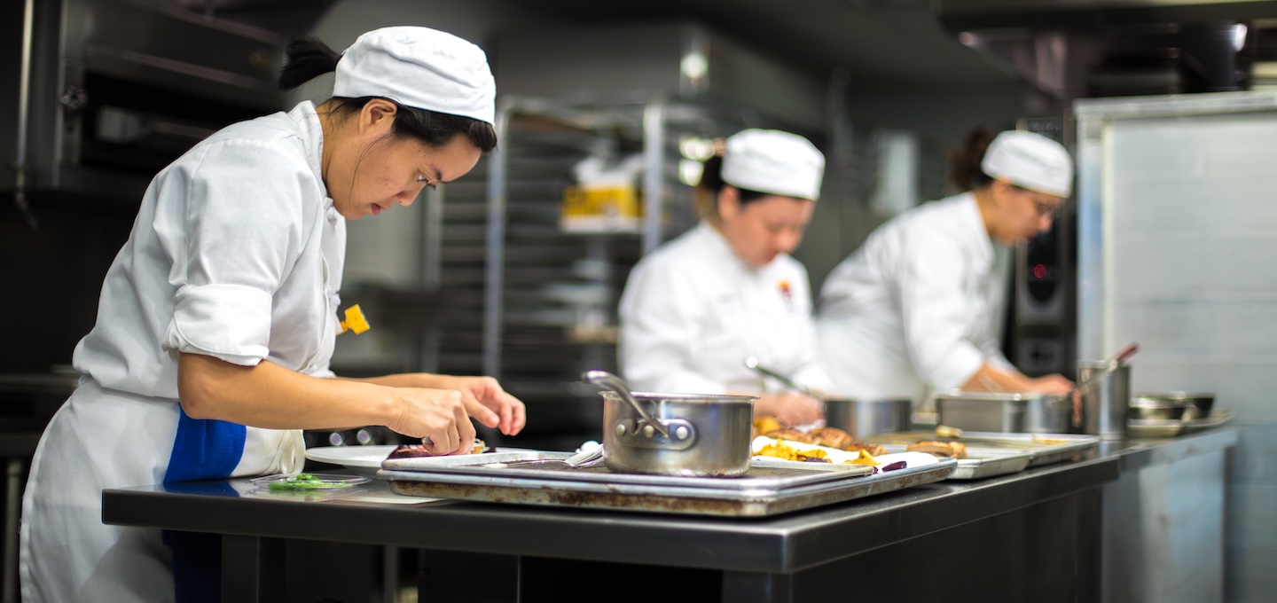Three students in chef whites plate dishes in a kitchen classroom