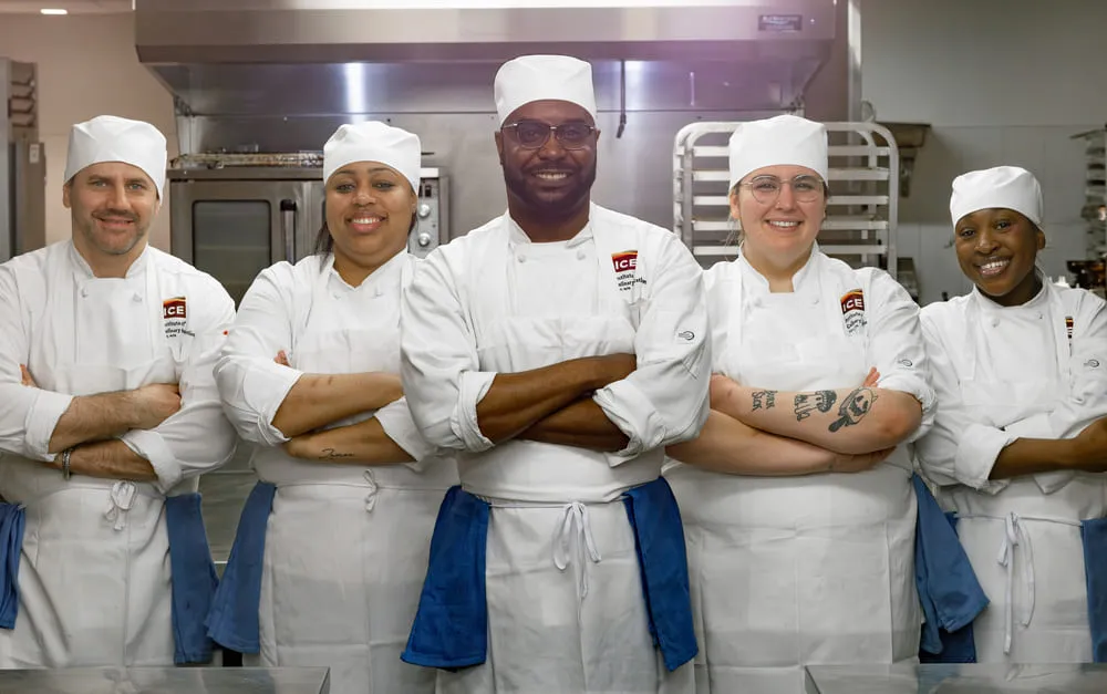 5 chefs lined up in professional kitchen wearing aprons and toques