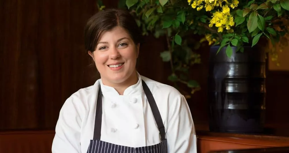 ICE 50th Anniversary Alumni Honoree Chef Michelle Palazzo wears chef whites and apron while smiling in front of a black vase with yellow flowers. 