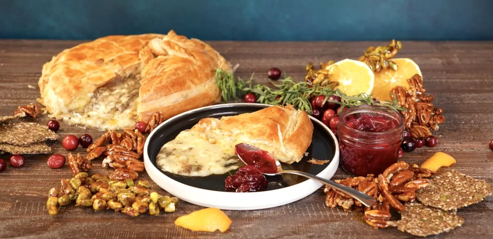 A slice of baked Brie cheese wrapped in baked puff pastry sits on a black plate in front of a wheel of the baked Brie cheese. They are both surrounded by green herbs, orange halves, candied nuts and cranberries