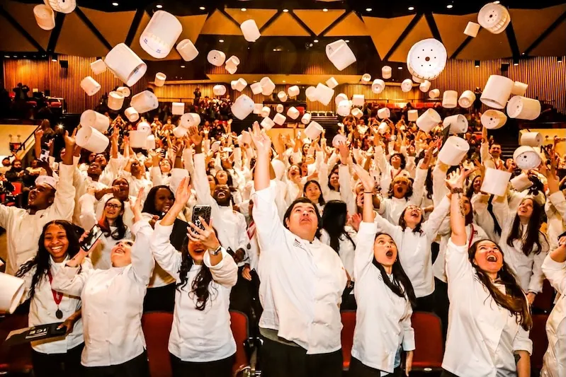 A large group of graduating culinary students wearing white chef coats celebrate inside an auditorium. They are cheering, smiling, and throwing their tall white chef hats (toques) high into the air.