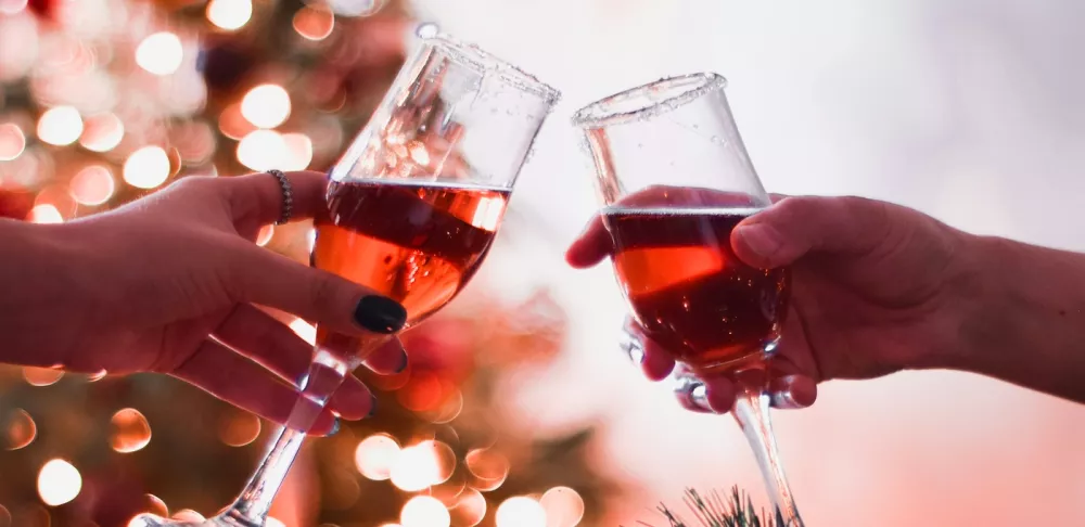 Two hands clink wine glasses with red liquid together above a table with garland and ornaments on it