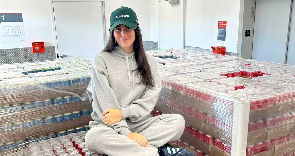 Danielle Krueger, a woman with long brown hair wearing a green hat that reads "Sunbear" and wearing a light grey sweat suit, sits on pallets of cans