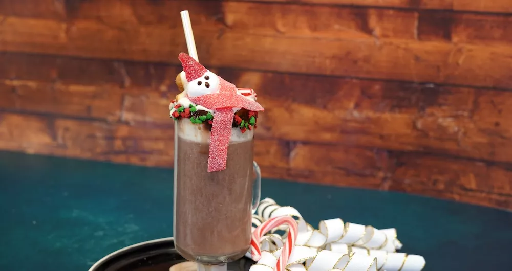 A clear mug of hot chocolate decorated with red and green holiday sprinkles and topped with a marshmallow shaped like a snowman wearing a red gumdrop hat