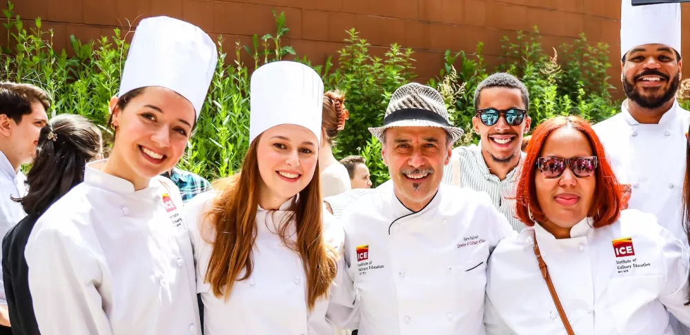 Multiple ICE graduates in white chef's coats and hats smile at ICE's commencement ceremony