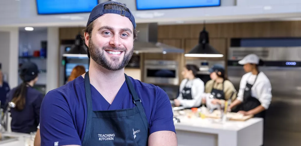 Dr. Nate Wood, a man with dark hair, blue eyes and a backwards blue baseball cap, smiles in a hospital teaching kitchen with his arms crossed