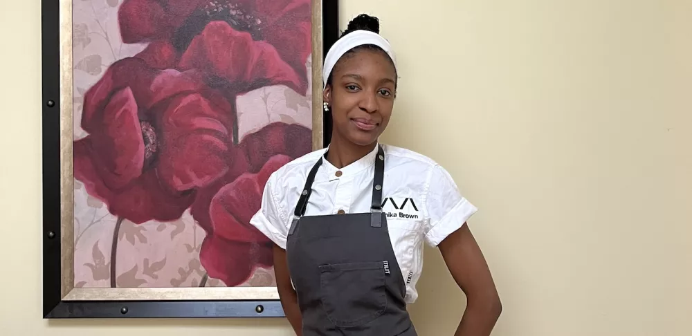 ICE graduate Onika Brown, a woman with black hair pulled back with a white headband wearing a white chef's coat and a grey apron, smiles in front of a framed painting of red flowers