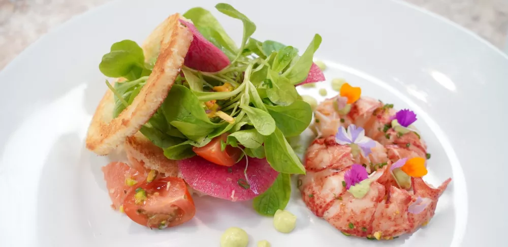 A green salad with colorful vegetables sits on a white plate next to red lobster