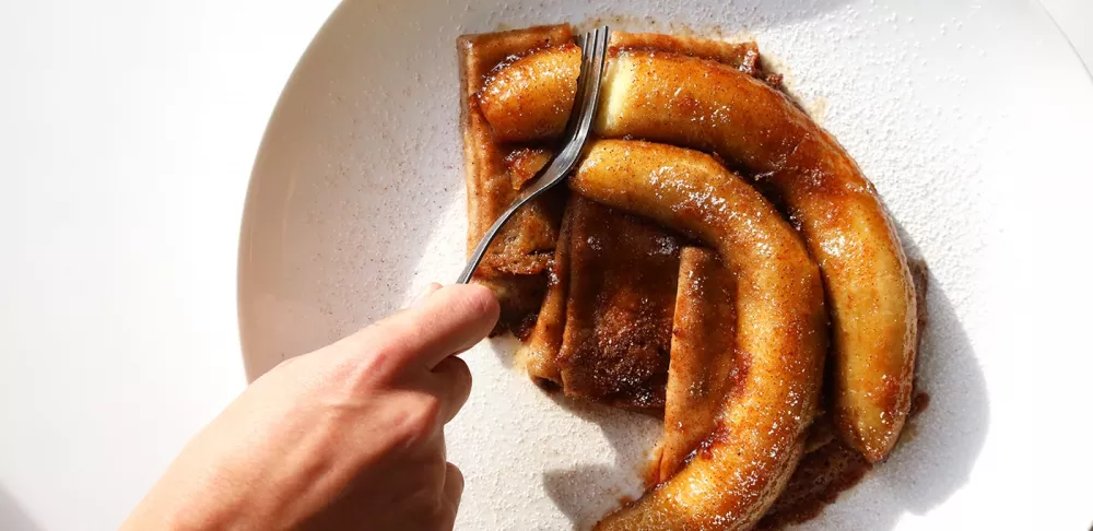 A hand cuts into crepes with caramelized bananas on top, all on a white plate