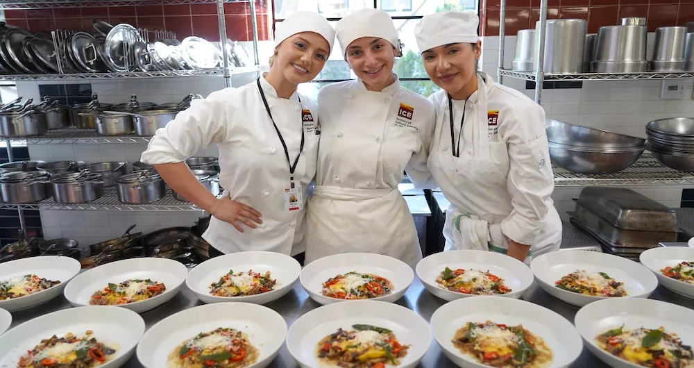 Three students in chef whites smile behind a table of plated meat dishes