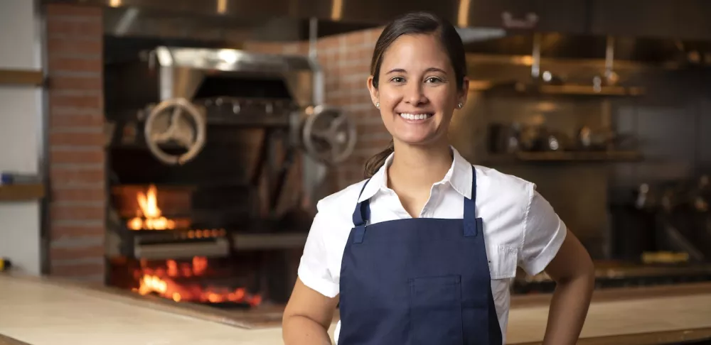 Suzanne Cupps, a brunette woman with her hair in a ponytail, wears a white collared shirt and a blue apron and smiles in front of an open kitchen with a wood-fired stove