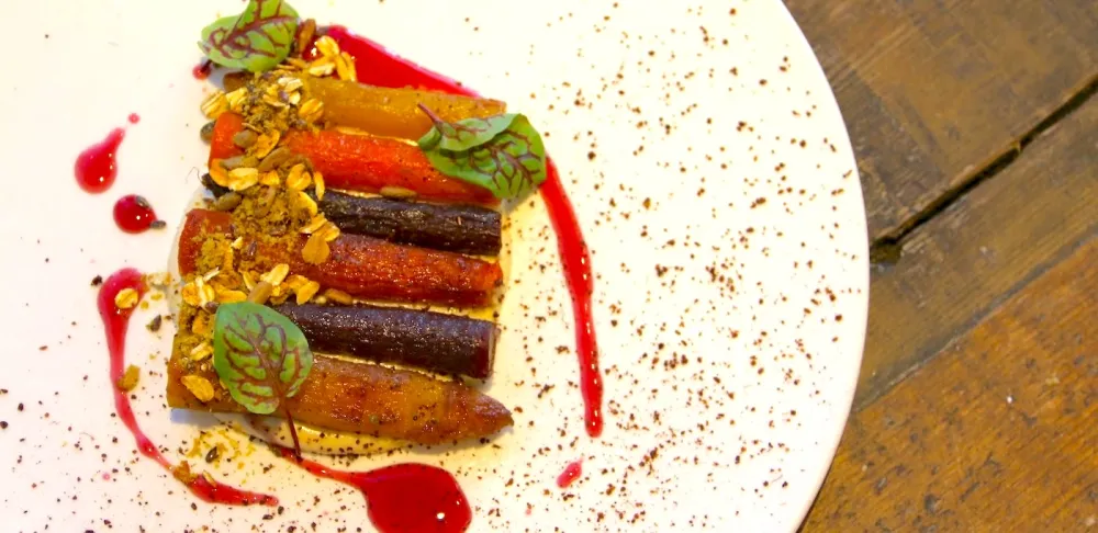 Roasted multi-colored carrots topped with green herbs and a crispy buckwheat crumble sit on a white plate