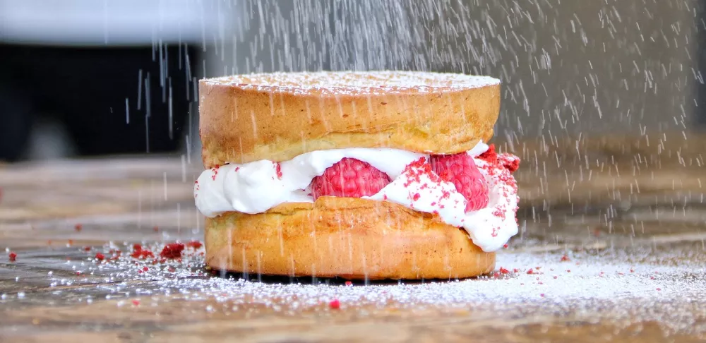 A choux pastry with white cream and raspberries dusted with powdered sugar sits on a brown board