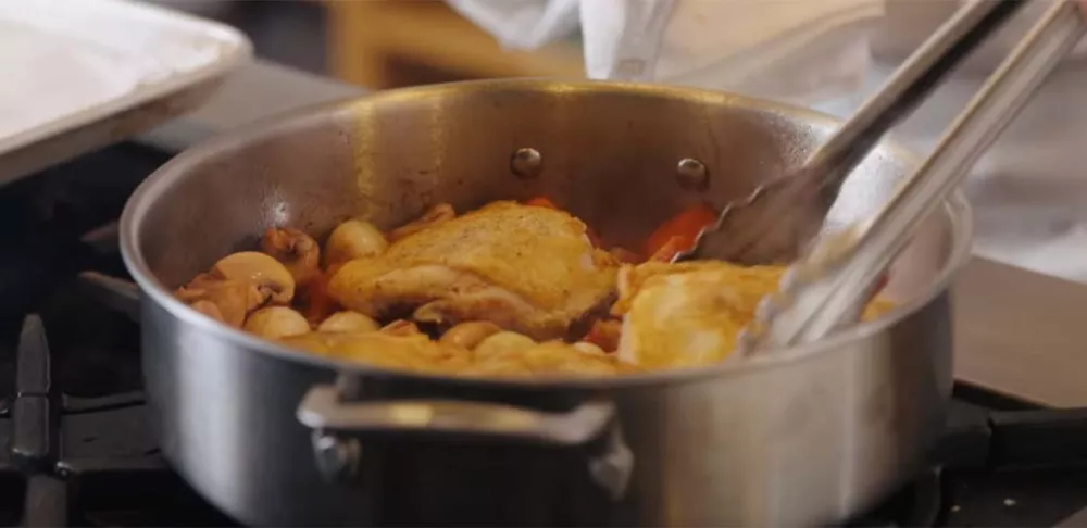 Tongs grab a piece of chicken being cooked as coq au vin in a steel pan