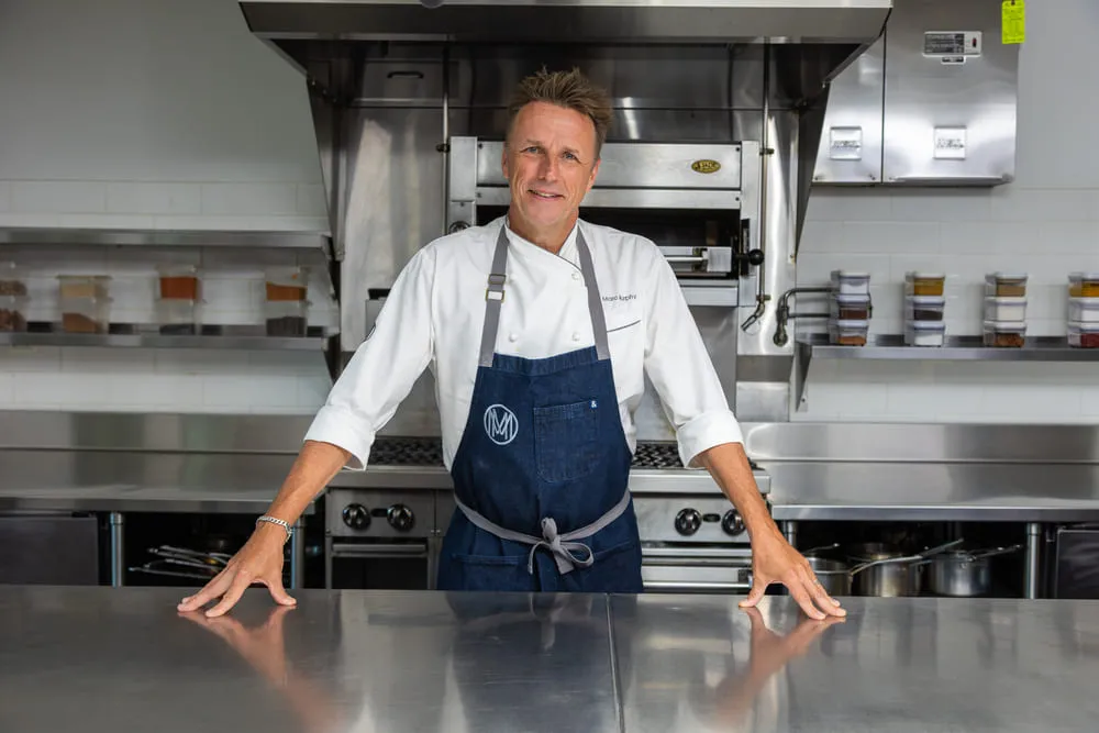 Chef Marc Murphy in chef whites and navy apron stands at counter in industrial kitchen.
