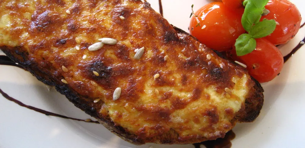 Cheesy toasted welsh rarebit sits on a white plate next to small tomatoes