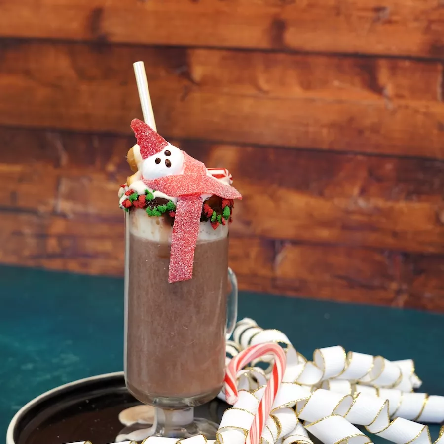 A clear mug of hot chocolate decorated with red and green holiday sprinkles and topped with a marshmallow shaped like a snowman wearing a red gumdrop hat