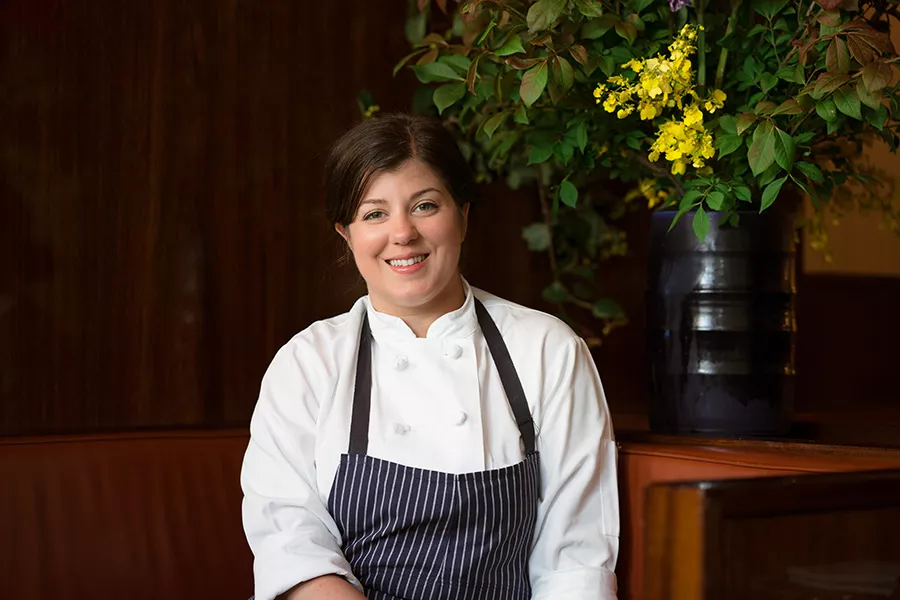 ICE 50th Anniversary Alumni Honoree Chef Michelle Palazzo wears chef whites and apron while smiling in front of a black vase with yellow flowers. 