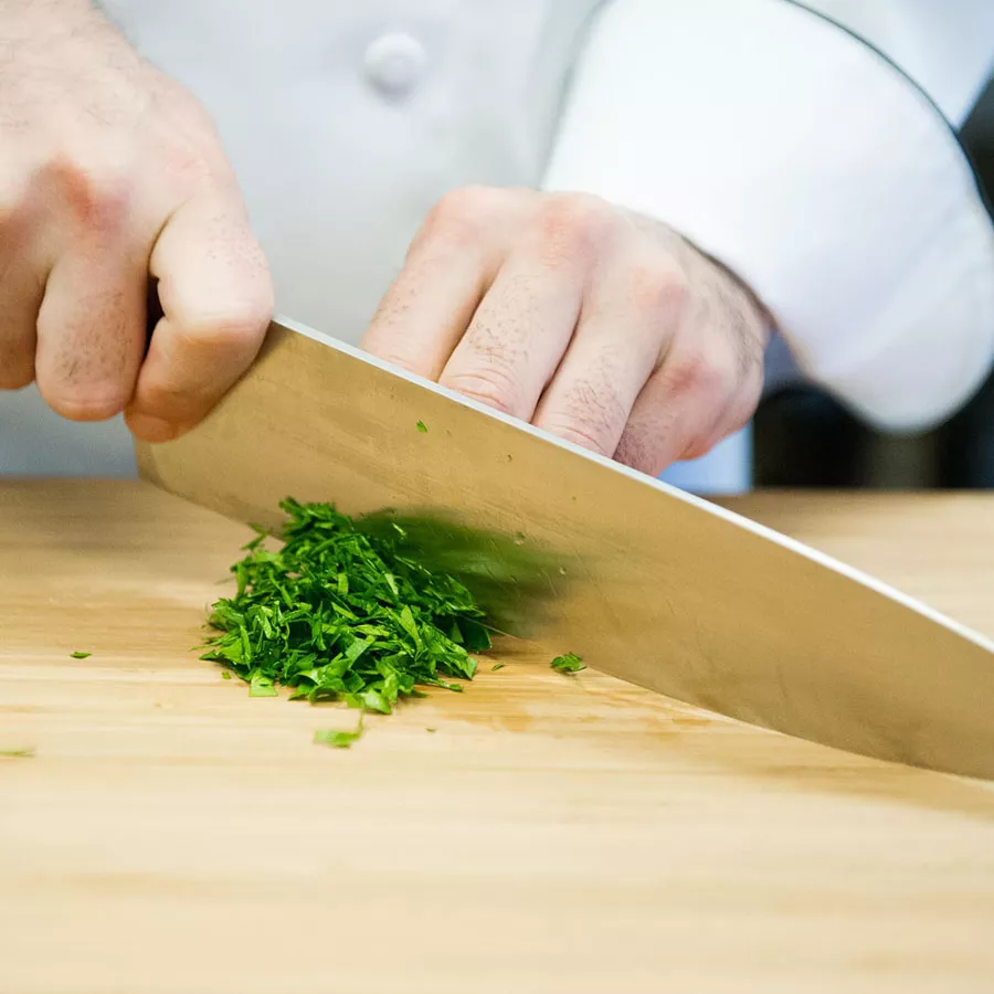 a chef's hands use a knife to cut cilantro on a wooden cutting board