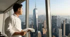 A chef in a white uniform holds a knife, gazing out a large window at the city skyline, featuring a prominent skyscraper under a clear sky.